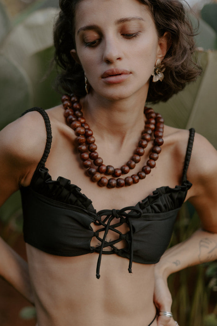A model wearing a black bikini top with ruffle detailing and a corset-style front tie, standing in front of lush greenery.